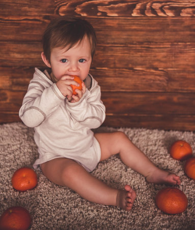 Cute baby boy is looking at camera and playing with mandarins while sitting on carpet at homeの写真素材