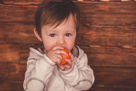 Cute baby boy is looking at camera and playing with mandarins while sitting on carpet at homeの写真素材