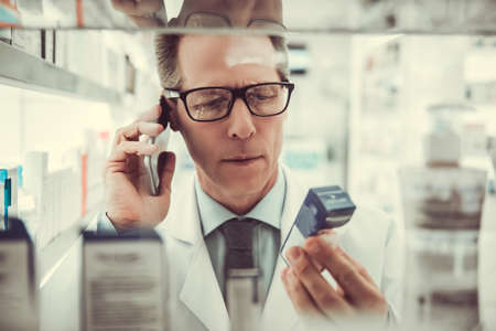 Handsome pharmacist is talking on the mobile phone and searching for a medication while working in pharmacyの写真素材