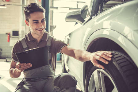 Handsome young auto mechanic in uniform is making notes and smiling while examining car in auto serviceの写真素材