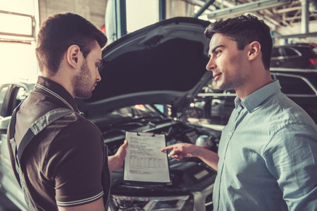 At the auto service. Handsome young auto mechanic in uniform is talking with a client and showing a document to fillの写真素材