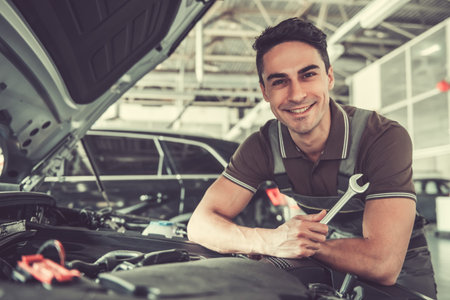 Handsome young auto mechanic in uniform is holding a spanner, looking at camera and smiling while repairing car in auto serviceの写真素材