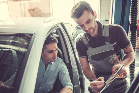 At the auto service. Handsome young auto mechanic in uniform is talking with a client and showing a document to fillの写真素材