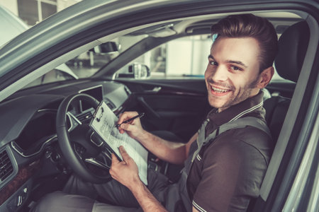 Handsome young auto mechanic in uniform is making notes, looking at camera and smiling while examining car in auto serviceの写真素材