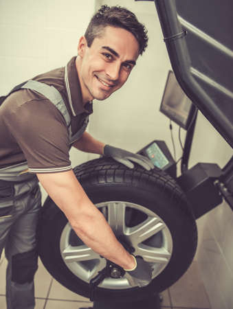 At the auto service. Handsome young auto mechanic in uniform is looking at camera and smiling while performing wheel diagnosisの写真素材