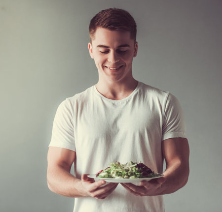 Handsome guy is smiling while holding a plate with salad, on gray backgroundの写真素材