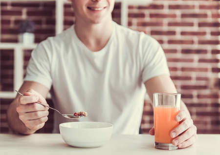 Handsome young guy is smiling while eating cereals in kitchen at homeの写真素材