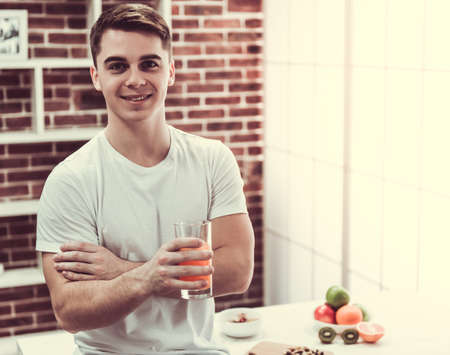 Handsome young guy is drinking orange juice, looking at camera and smiling while standing in kitchen at homeの写真素材