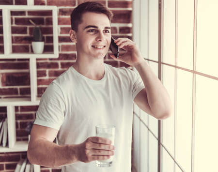 Handsome young guy is drinking milk, talking on the mobile phone and smiling while standing in kitchen at homeの写真素材