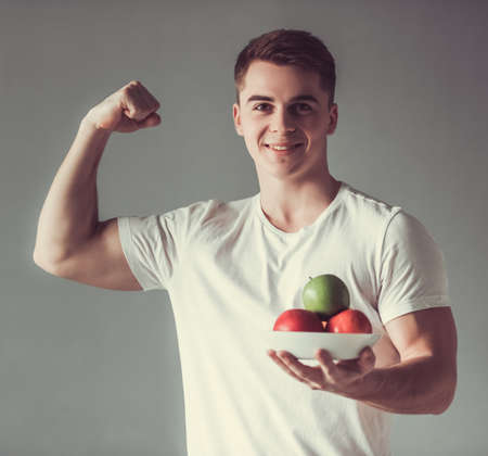 Handsome guy is holding fruits, showing his muscles, looking at camera and smiling, on gray backgroundの写真素材