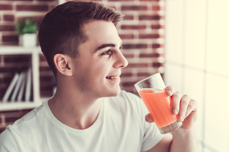 Handsome young guy is smiling while drinking orange juice in kitchen at homeの写真素材