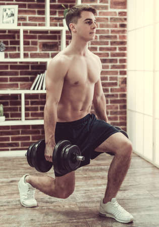 Handsome muscular guy with bare torso is doing lunges with dumbbells while working out at homeの写真素材