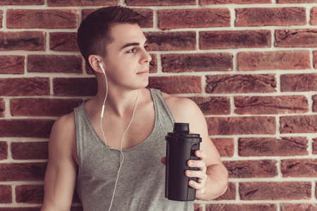 Handsome muscular guy in earphones is  listening to music and holding a shaker, on brick wall backgroundの写真素材