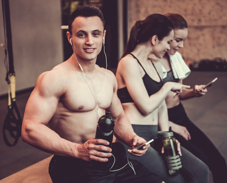 Beautiful young sports people are resting after workout in gym. Guy is looking at camera and smiling