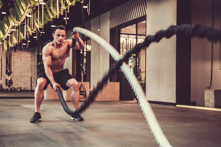 Handsome muscular man is doing battle rope exercise while working out in gymの写真素材