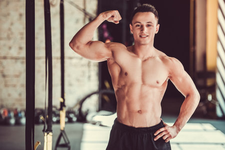 Handsome young muscular sportsman is showing his muscles, looking at camera and smiling while...