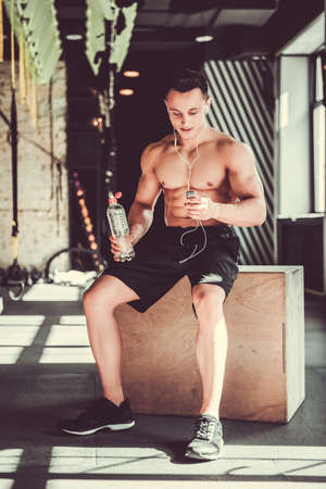 Handsome young muscular sportsman is listening to music, holding a bottle of water and smiling after workout in gymの写真素材