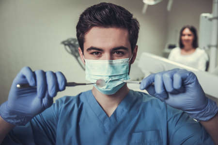 Handsome young dentist in surgical mask is holding tools and looking at cameraの写真素材
