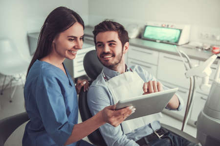 Handsome young patient is sitting in chair while visiting a beautiful female dentist. Both are using a digital tablet and smilingの写真素材