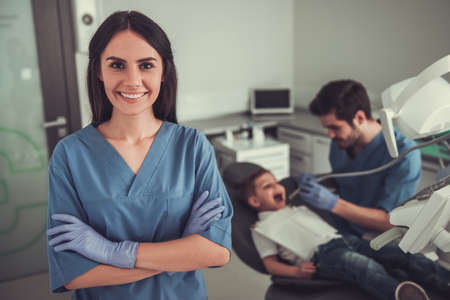 Beautiful young female dentist is looking at camera and smiling, little boy visiting dentist in the backgroundの写真素材