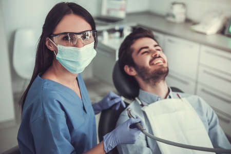 Handsome young patient is sitting in chair and smiling while visiting a beautiful female dentistの写真素材