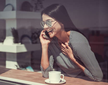 The view through the window. The young beautiful girl drinking coffee while sitting in a cafe and emotionally talking on the phoneの写真素材