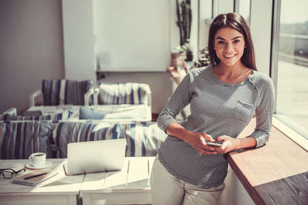 Cheerful young pretty girl using phone standing near a large window in the cafe.の写真素材