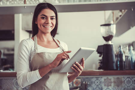 Wellcome to my cafe. Portrait of a young girl cheerful barista ready to receive orders.の写真素材