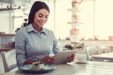The young beautiful girl drinking coffee and dessert eating, sitting in a cafe on the terrace. Using an electronic tablet.の写真素材