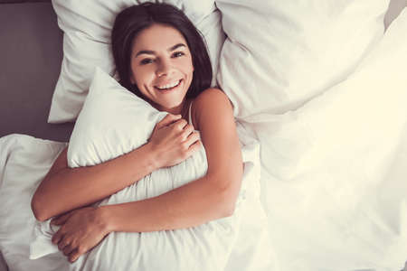 Young beautiful girl smiling while lying in bed hugging pillow top viewの写真素材