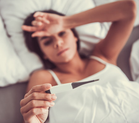 Young beautiful woman lying in bed, she measures her temperature with a thermometer and touching her foreheadの写真素材
