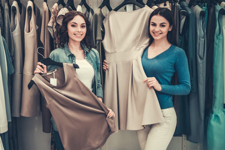 Beautiful girls are choosing clothes, looking at camera and smiling while doing shopping in boutique