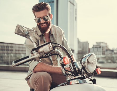 Handsome bearded guy in sun glasses is looking at camera and smiling while sitting on his scooterの写真素材