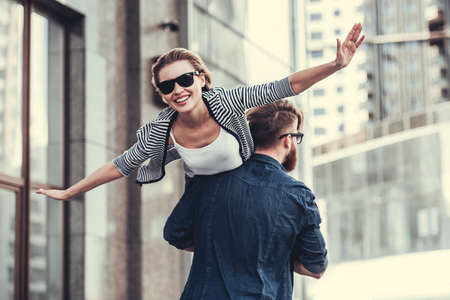 Beautiful young couple in sun glasses is walking down the street. Guy is carrying girl in his arms, she is smilingの写真素材