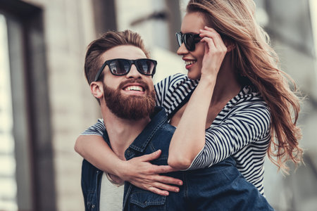 Beautiful young couple in sun glasses looking at each other and smiling while standing outdoors. Girl piggybackの写真素材