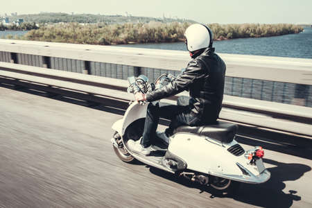 Guy in leather jacket and helmet is riding on scooter through the city bridgeの写真素材