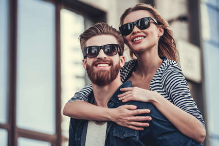 Beautiful young couple in sun glasses looking away and smiling while standing outdoors. Girl piggybackの写真素材