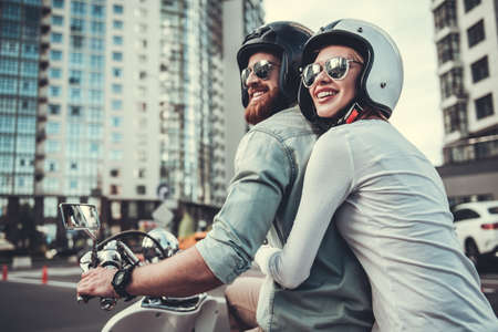 Beautiful young couple in sun glasses and helmets is smiling while riding a scooterの写真素材