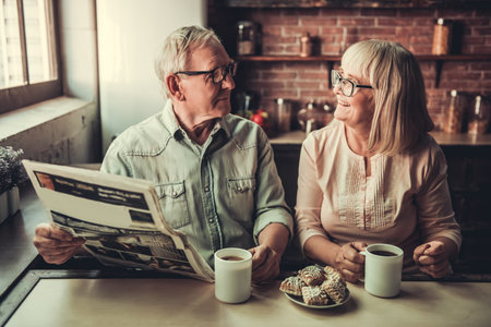 Beautiful senior couple is drinking tea, reading newspaper and smiling while resting in kitchenの写真素材