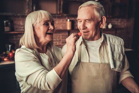 Beautiful senior couple in aprons is tasting food and smiling while cooking together in kitchenの写真素材