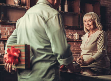 Beautiful senior woman is cooking and smiling while her husband is holding a present for her behind his backの写真素材
