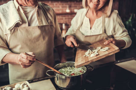 Beautiful senior couple in aprons is talking and smiling while cooking together in kitchenの写真素材