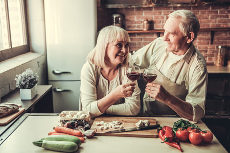 Beautiful senior couple in aprons is drinking wine, talking and smiling while cooking together in kitchenの写真素材