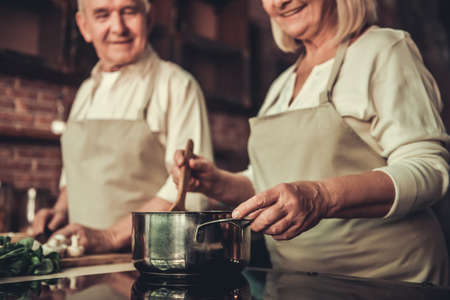 Beautiful senior couple in aprons is talking and smiling while cooking together in kitchenの写真素材