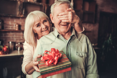 Beautiful senior woman is covering her husband eyes smiling while giving him a presentの写真素材