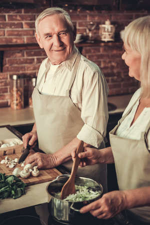 Beautiful senior couple in aprons is talking and smiling while cooking together in kitchenの写真素材
