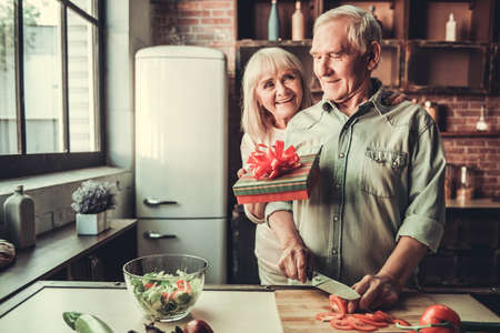 Handsome senior man is cooking and smiling while his wife is giving him a presentの写真素材