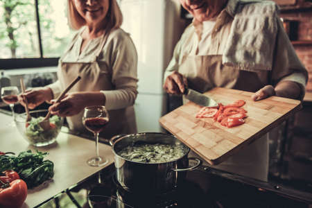 Beautiful senior couple in aprons is talking and smiling while cooking together in kitchenの写真素材
