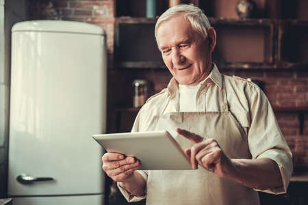 Handsome senior man in apron is using a digital tablet and smiling while cooking in kitchenの写真素材