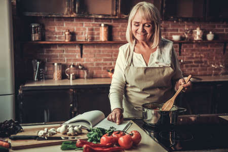 Beautiful senior woman in apron is reading a book of recipes and smiling while cooking in kitchenの写真素材